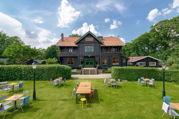 Un edificio grande de color oscuro con techo de tejas rojas, puerta verde y múltiples ventanas con detalles en verde. Frente al edificio, hay un césped bien cuidado con varias mesas y sillas dispuestas para comer al aire libre.