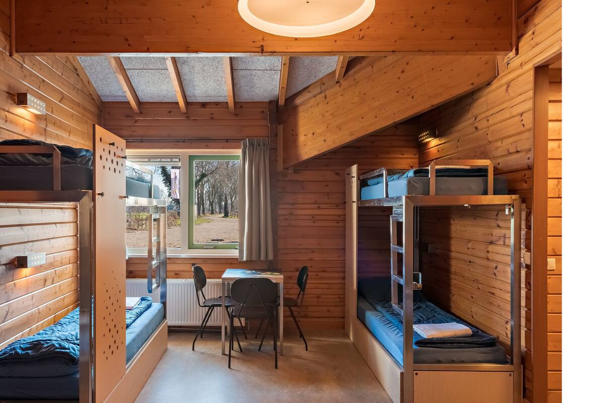 Una cabaña pequeña con literas, una mesita, sillas y una ventana con vistas a árboles.