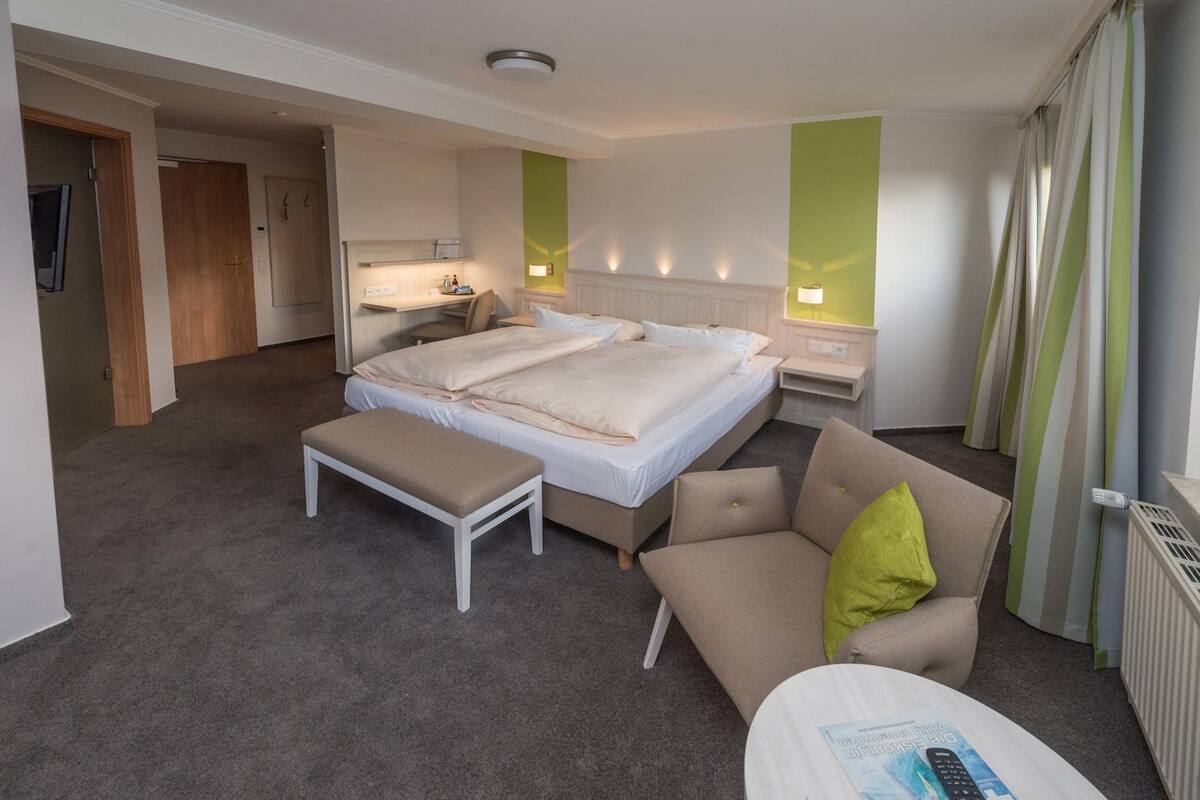 A hotel room with a large bed, a bedside table, a chair, and a small desk with a lamp.