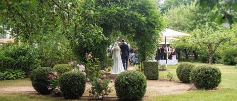 Un salón de bodas en un jardín con un pasillo central, rodeado de setos recortados y arbustos florecientes.