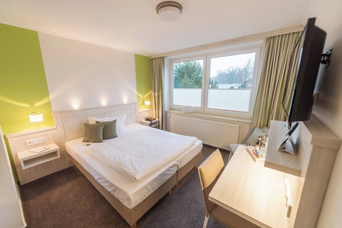 A hotel room with a bed, bedside tables, a desk, a chair, a TV, and a window with curtains.
