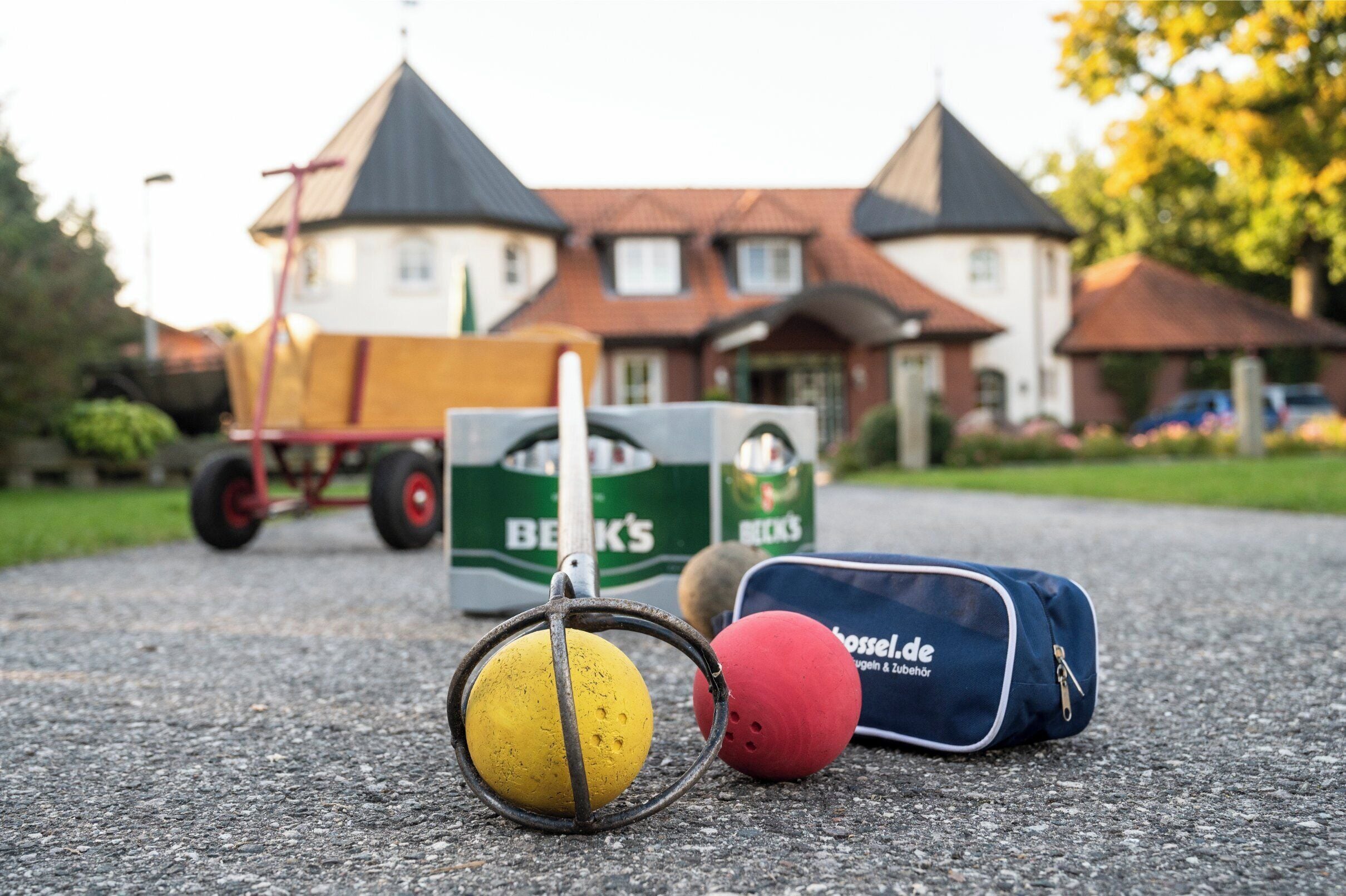 Ein Croquetball, ein Schläger und eine Tasche auf einer gepflasterten Fläche, im Hintergrund ein Haus und ein Wagen.
