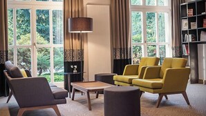 Een moderne woonkamer met een bank, fauteuil, salontafel, grote ramen met gordijnen en een boekenkast.