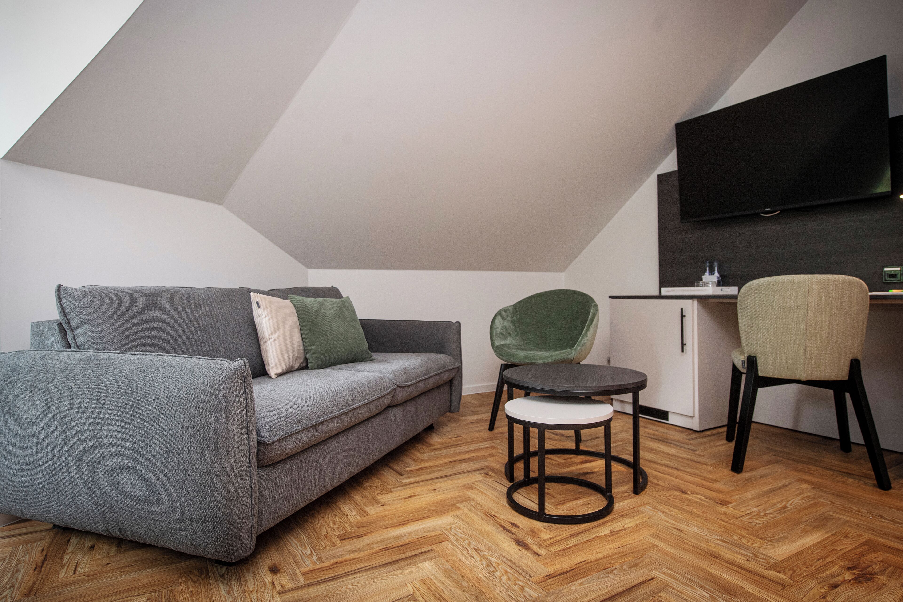 A modern loft bedroom with a sloped ceiling, a grey sofa, a green chair, a small round table, and a TV mounted on the wall.