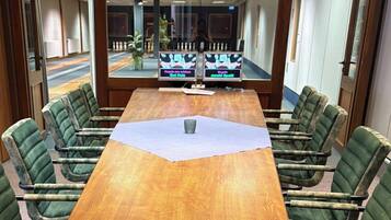 A conference room with a wooden table, green upholstered chairs, and two monitors displaying text.