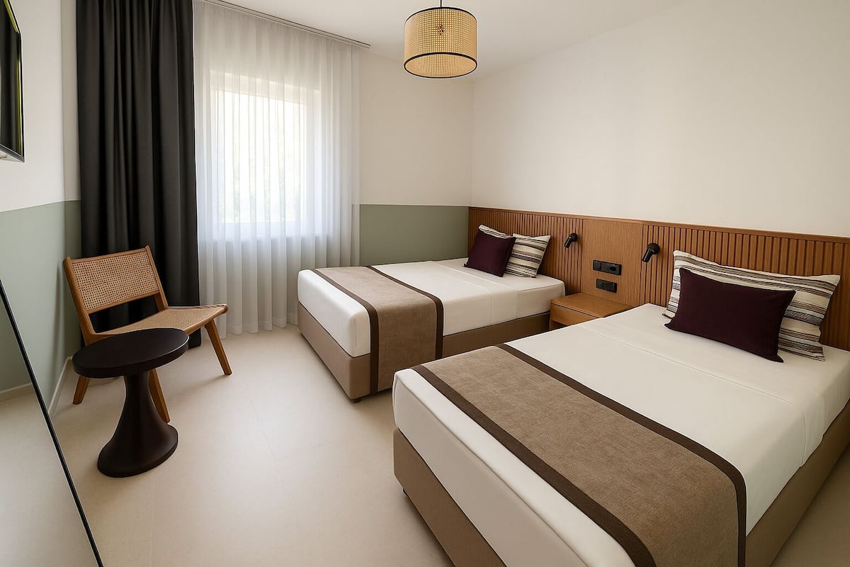 Hotellihuone, jossa on kaksi sänkyä, tuoli, pieni pöytä ja ikkuna, jossa on verhot.