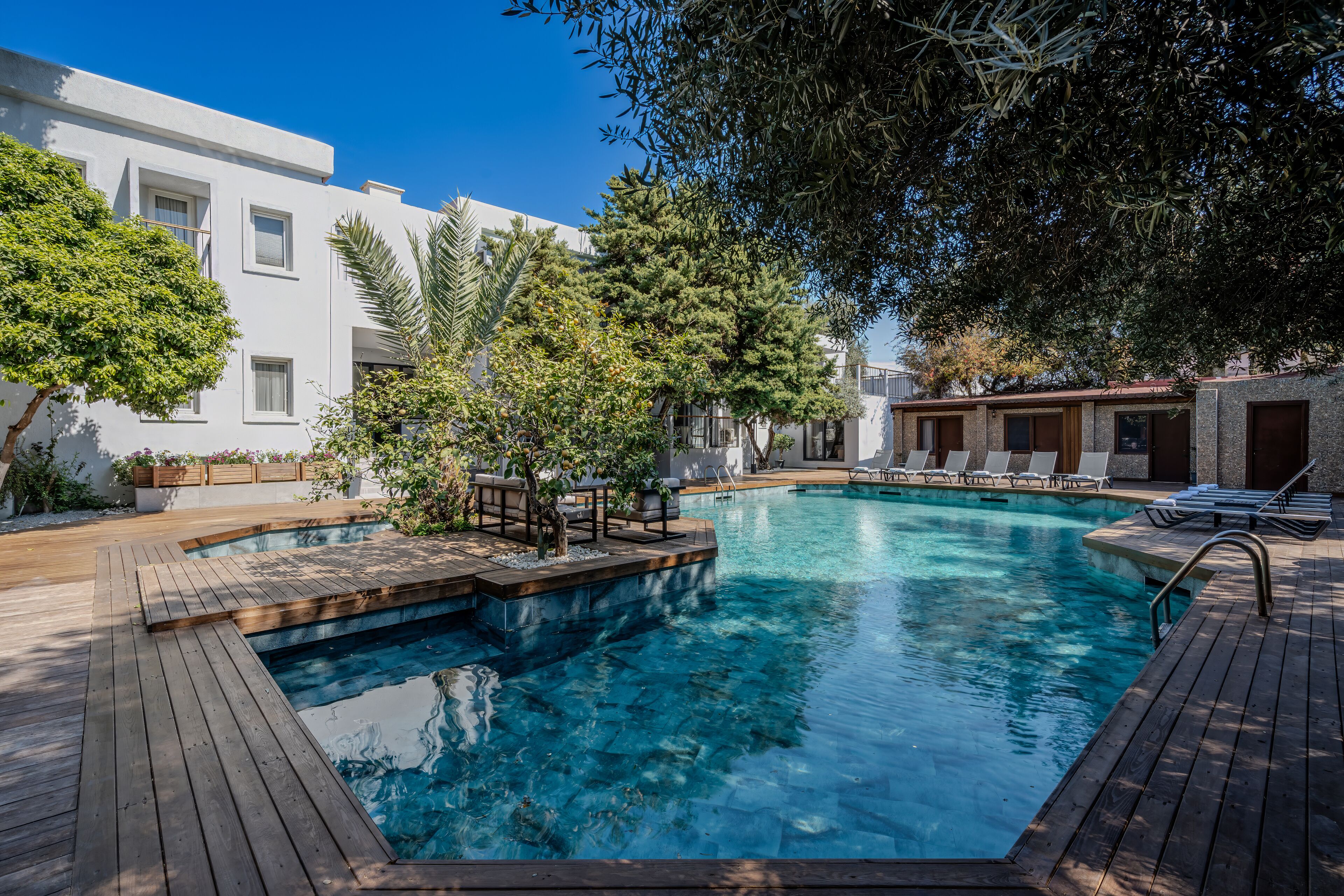 A swimming pool with wooden deck, lounge chairs, and a white building in the background.