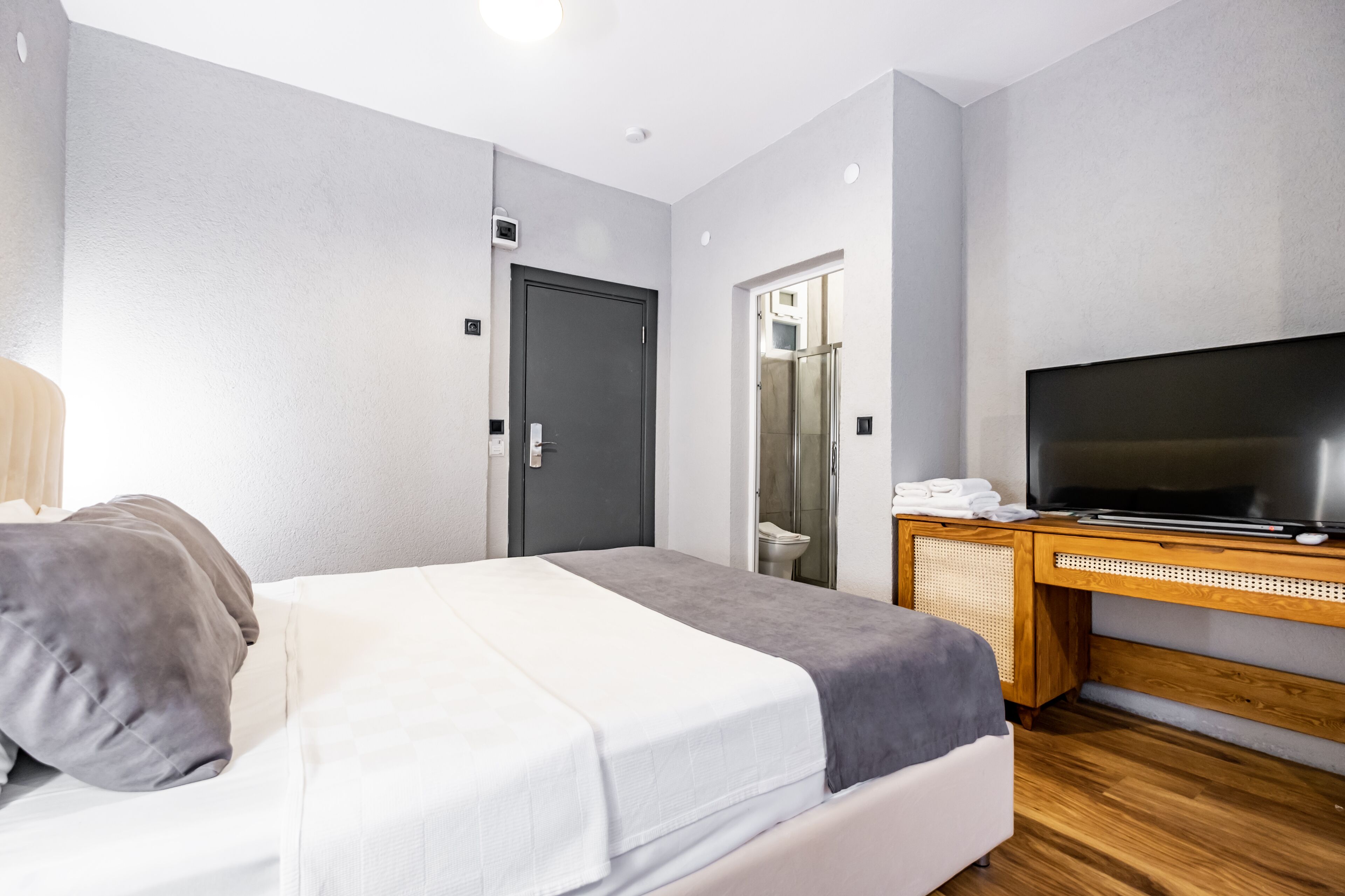 Quarto de hotel moderno com cama, televisão, escrivaninha de madeira e banheiro visível por uma porta aberta.