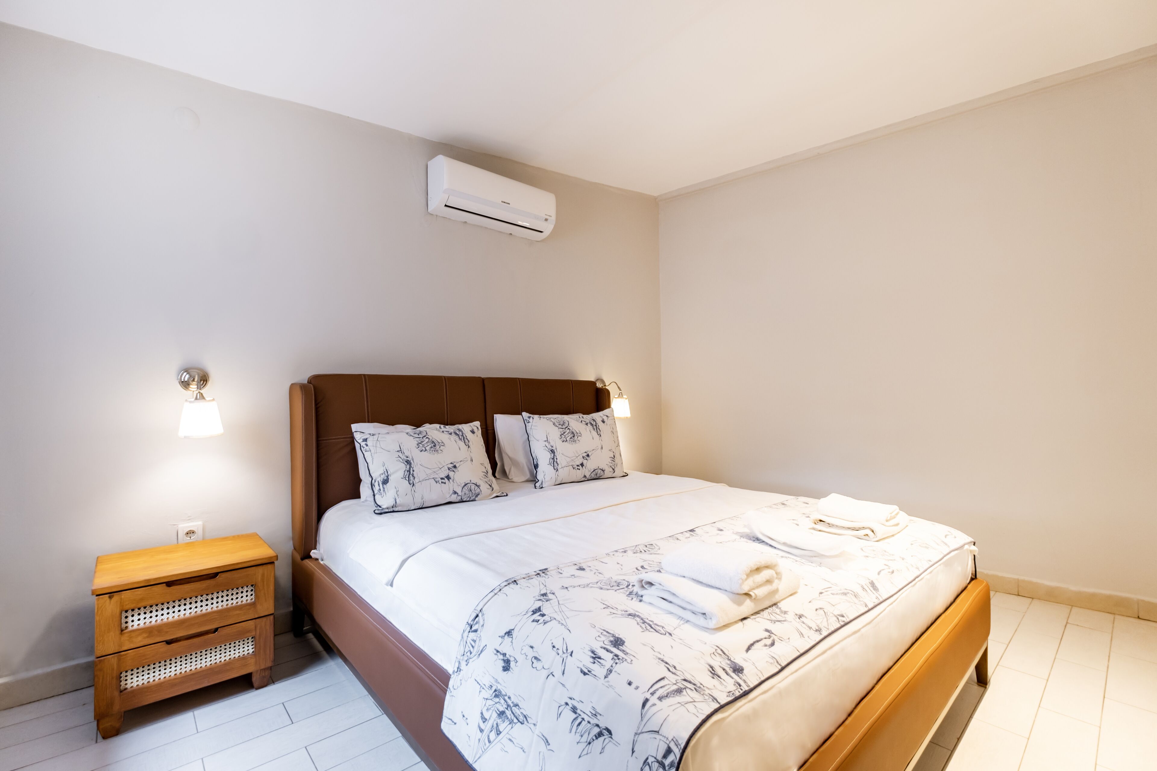 Una cama bien hecha con una colcha floral, una mesita de noche de madera con cajones y un aire acondicionado de pared.