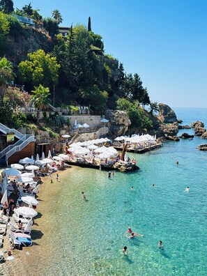 A coastal beach with clear water, sun loungers, umbrellas, and a rocky shoreline.