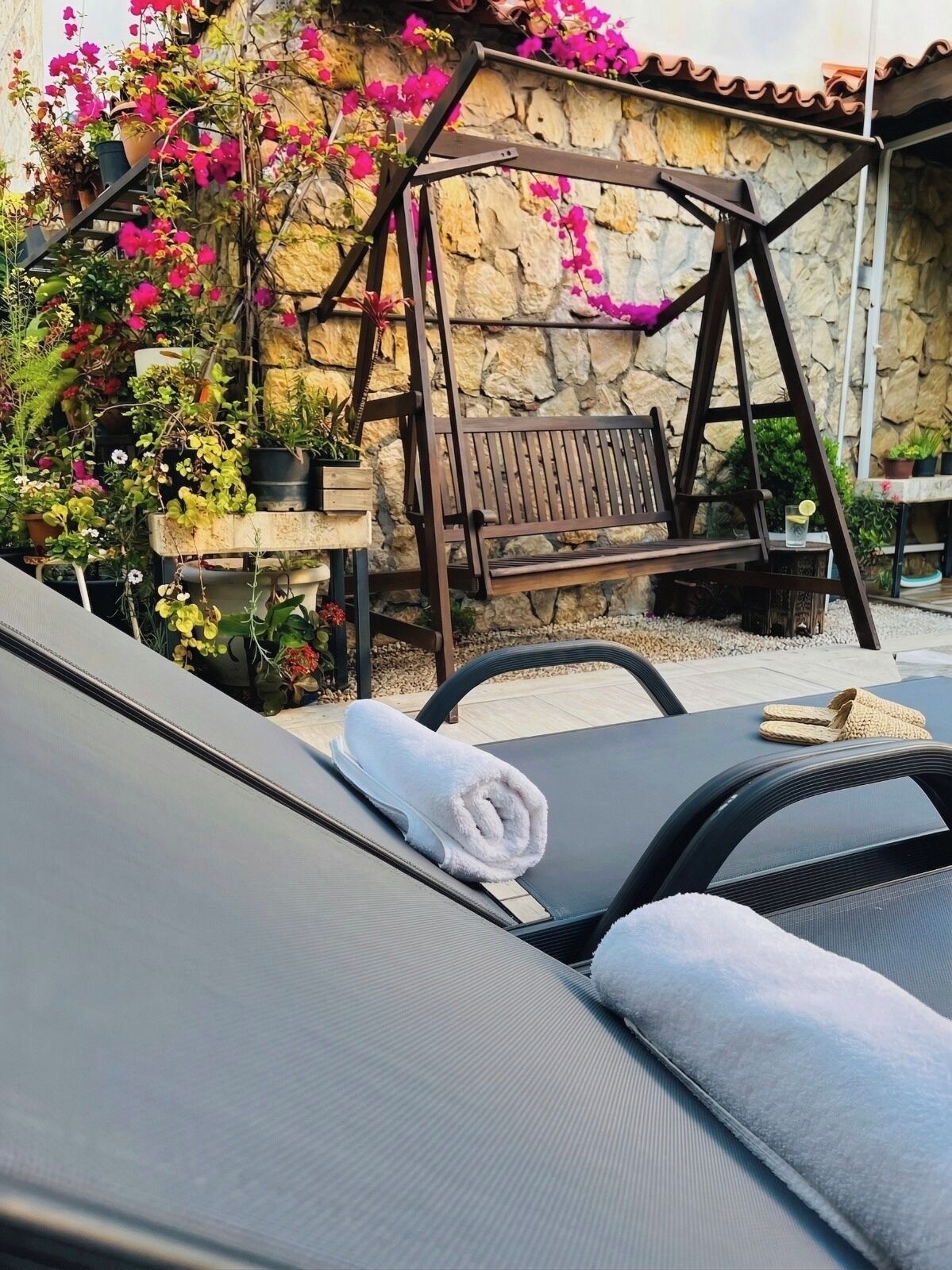 A sun lounger with a towel, a wooden swing, and potted plants against a stone wall.