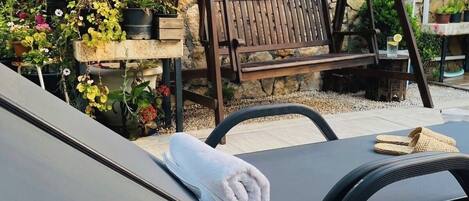 A sun lounger with a towel, a wooden swing, and potted plants against a stone wall.