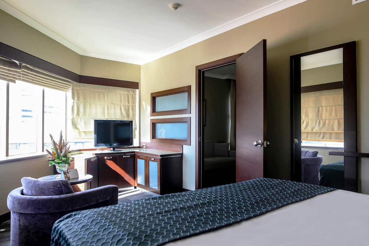 A hotel room with a bed, a desk with a television, a small seating area, and a window with blinds.