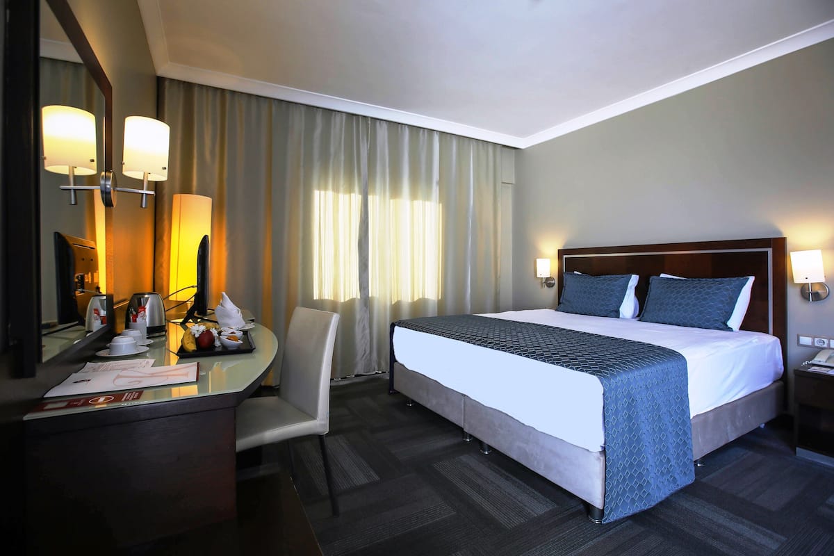 A hotel room with a large bed, a desk with a chair, a mirror, and a nightstand with a lamp.