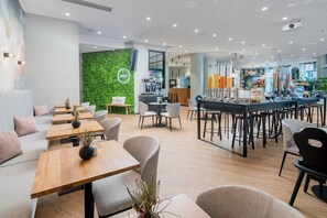 Un café moderne avec des tables et des chaises en bois, un mur végétal orné d’un logo et un coin bar avec des tabourets.