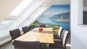 A dining area with a high table and chairs, a view of a lake and mountains through a large window, and a skylight above. - City Aparthotel Muenchen (Muenchen)