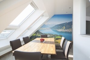 A dining area with a high table and chairs, a view of a lake and mountains through a large window, and a skylight above. - City Aparthotel Muenchen (Muenchen)