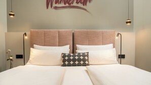 A neatly made bed with white linens and a patterned pillow, a wall-mounted sign reading "Wanderbar," and two pendant lights. - City Aparthotel Muenchen (Muenchen)
