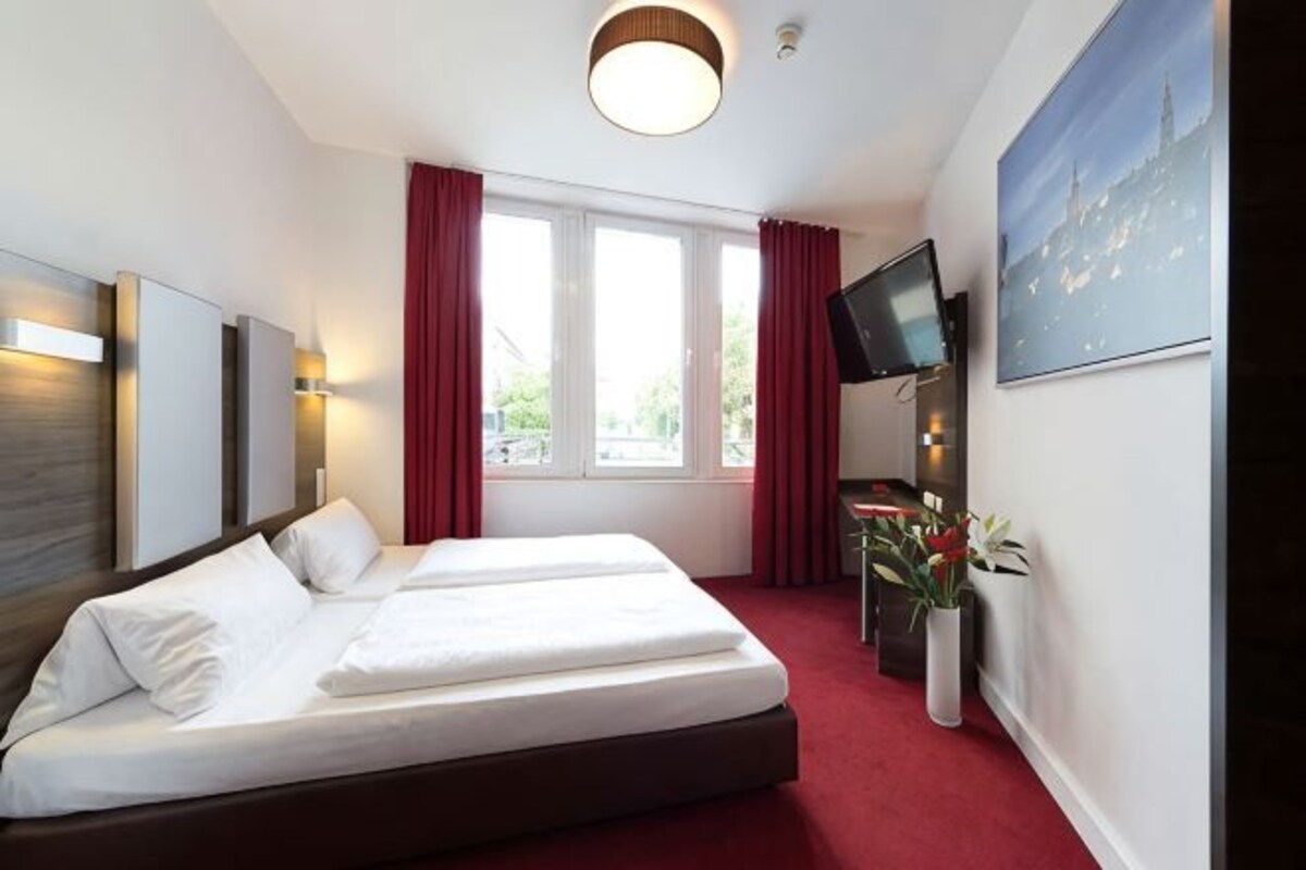 A hotel room with a bed, a TV, a desk, and a window with curtains.