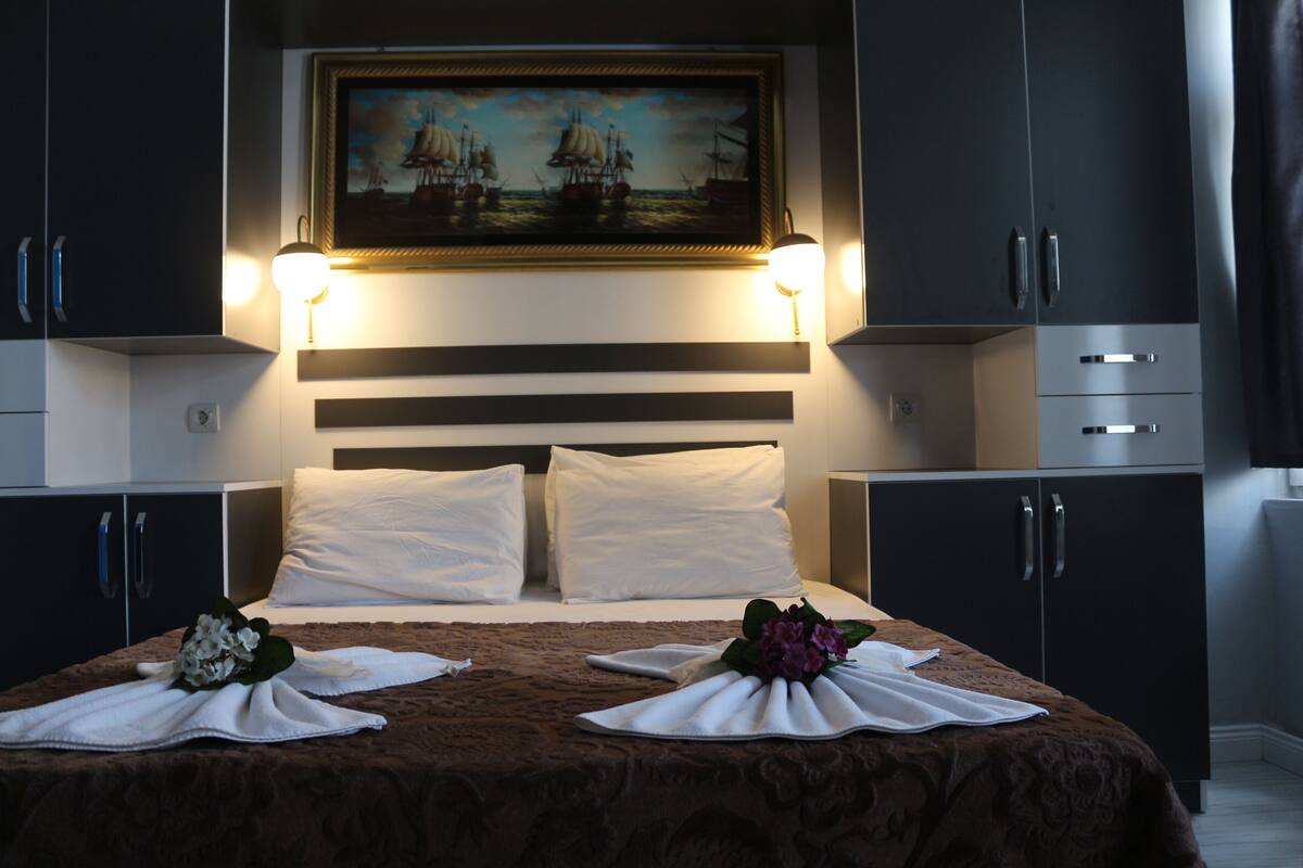 A neatly made bed with white linens and a brown bedspread, flanked by two nightstands with lamps, in a modern room with dark cabinetry and a framed painting above the bed.