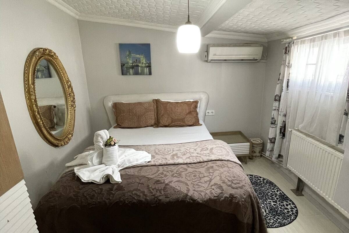 A hotel room with a bed, a mirror, a picture of a bridge, an air conditioner, and a window with curtains.