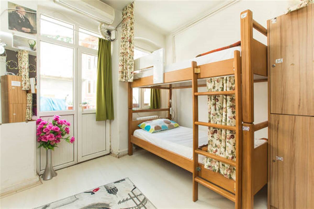 A bunk bed room with a wooden bunk bed, a small cabinet, a window with curtains, and a vase with flowers.