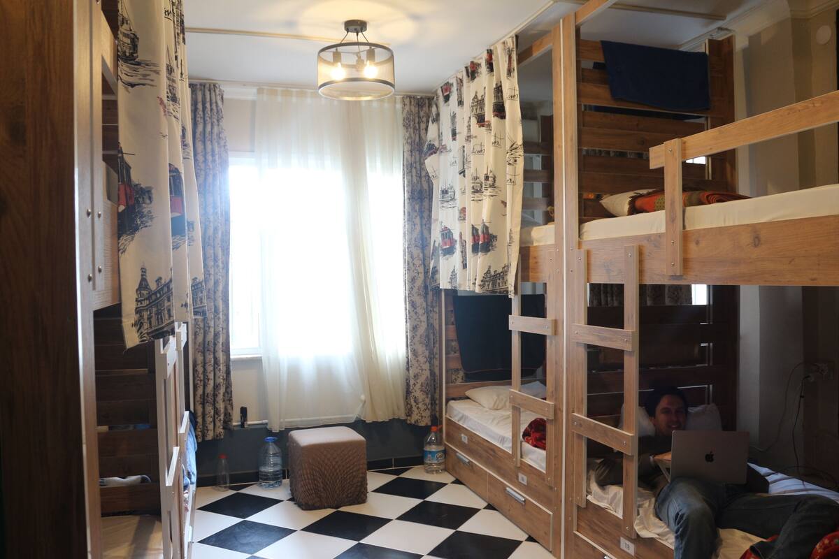 A dormitory room with bunk beds, a checkered floor, a window with curtains, and a ceiling light.