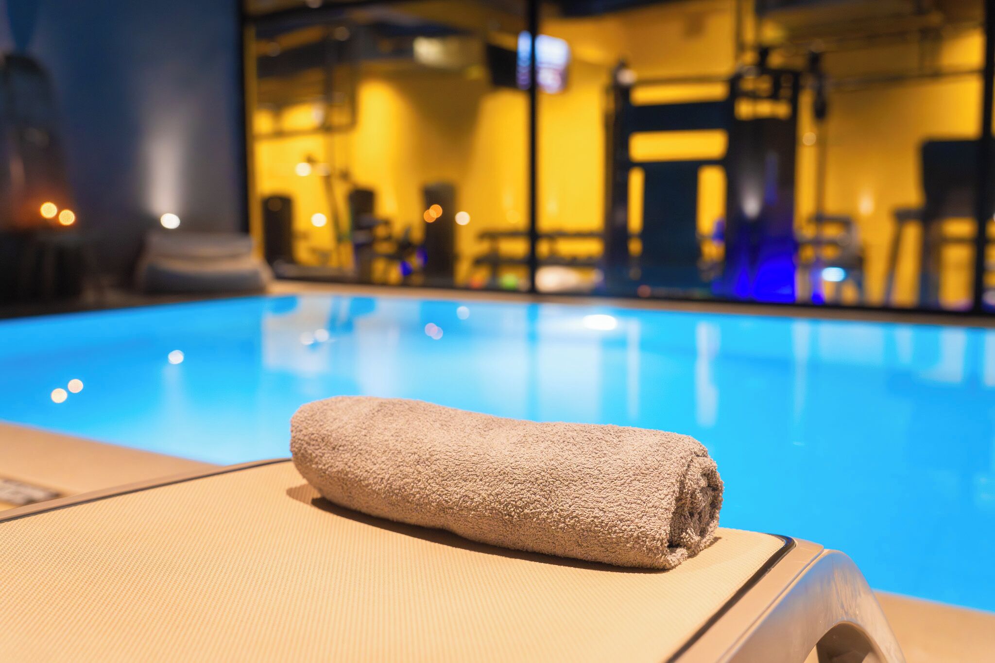 Ein Poolbereich mit einem Handtuch auf einem Liegestuhl, einer Glasbarriere und einem Raum mit gelber Beleuchtung im Hintergrund.