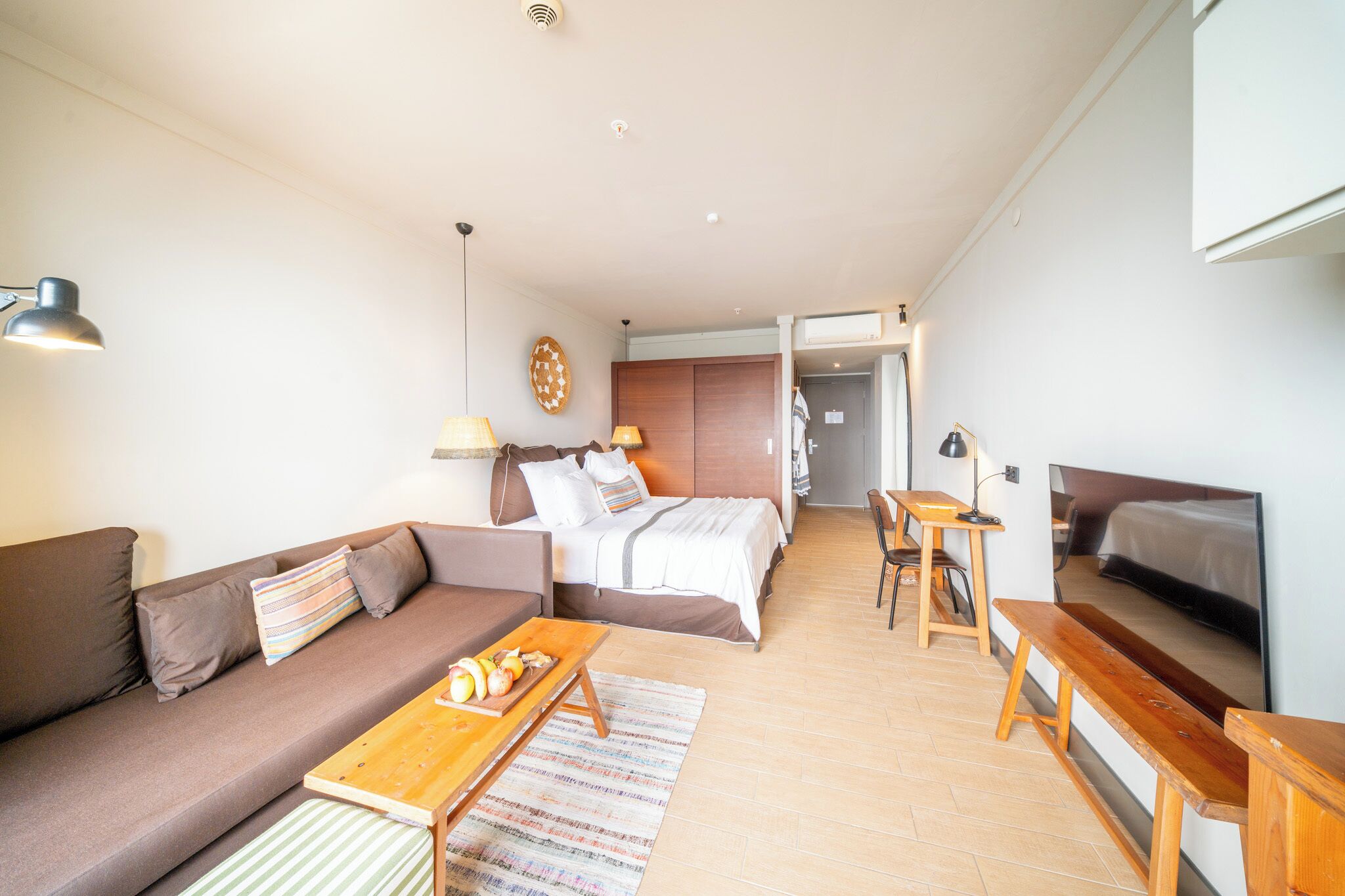 Une chambre d’hôtel moderne avec un canapé, une table en bois sur laquelle sont disposés des fruits, un lit, une télévision et un bureau.
