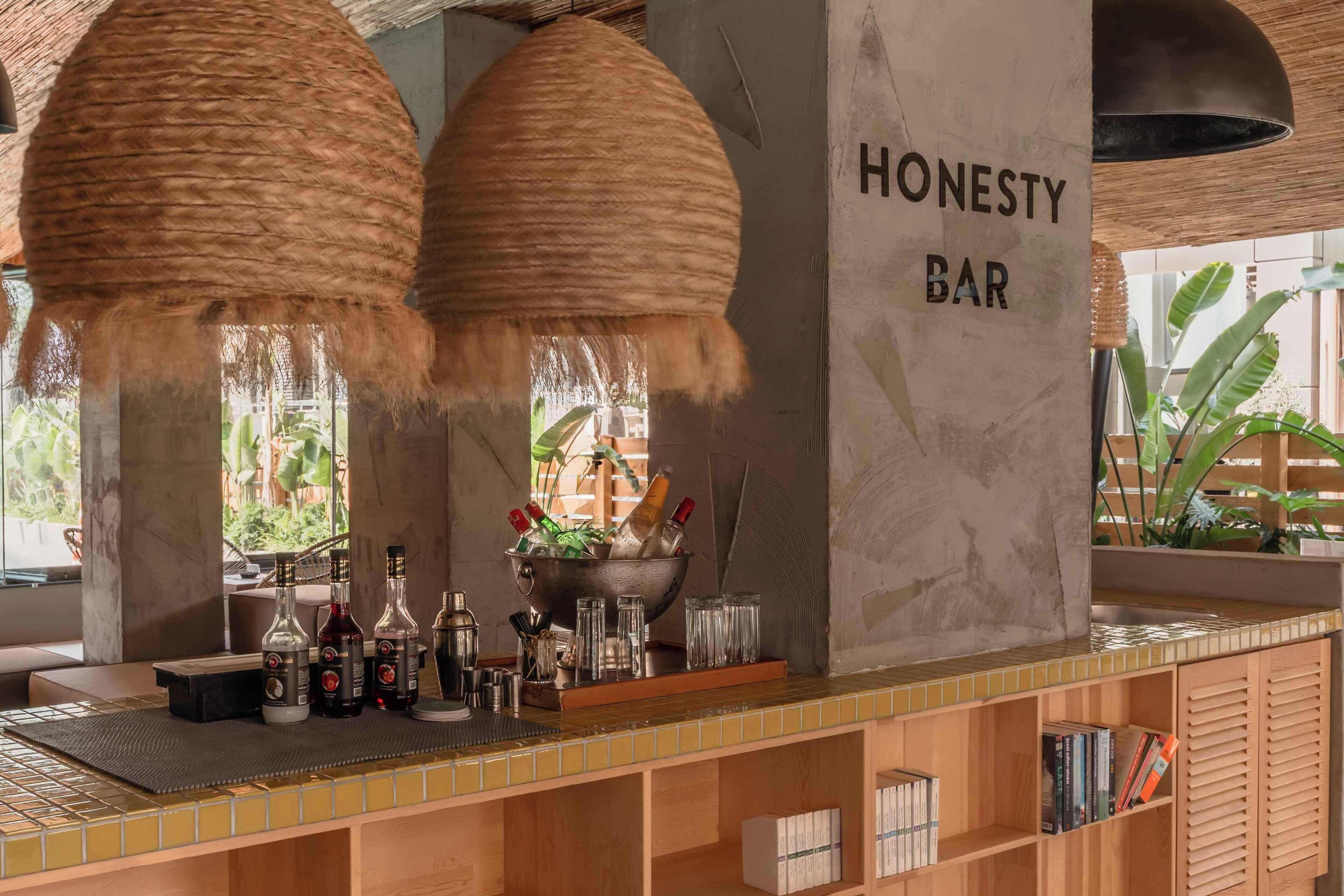 A bar area with a sign reading "HONESTY BAR," featuring a tiled counter, hanging woven lamps, and a variety of bottles and glasses.