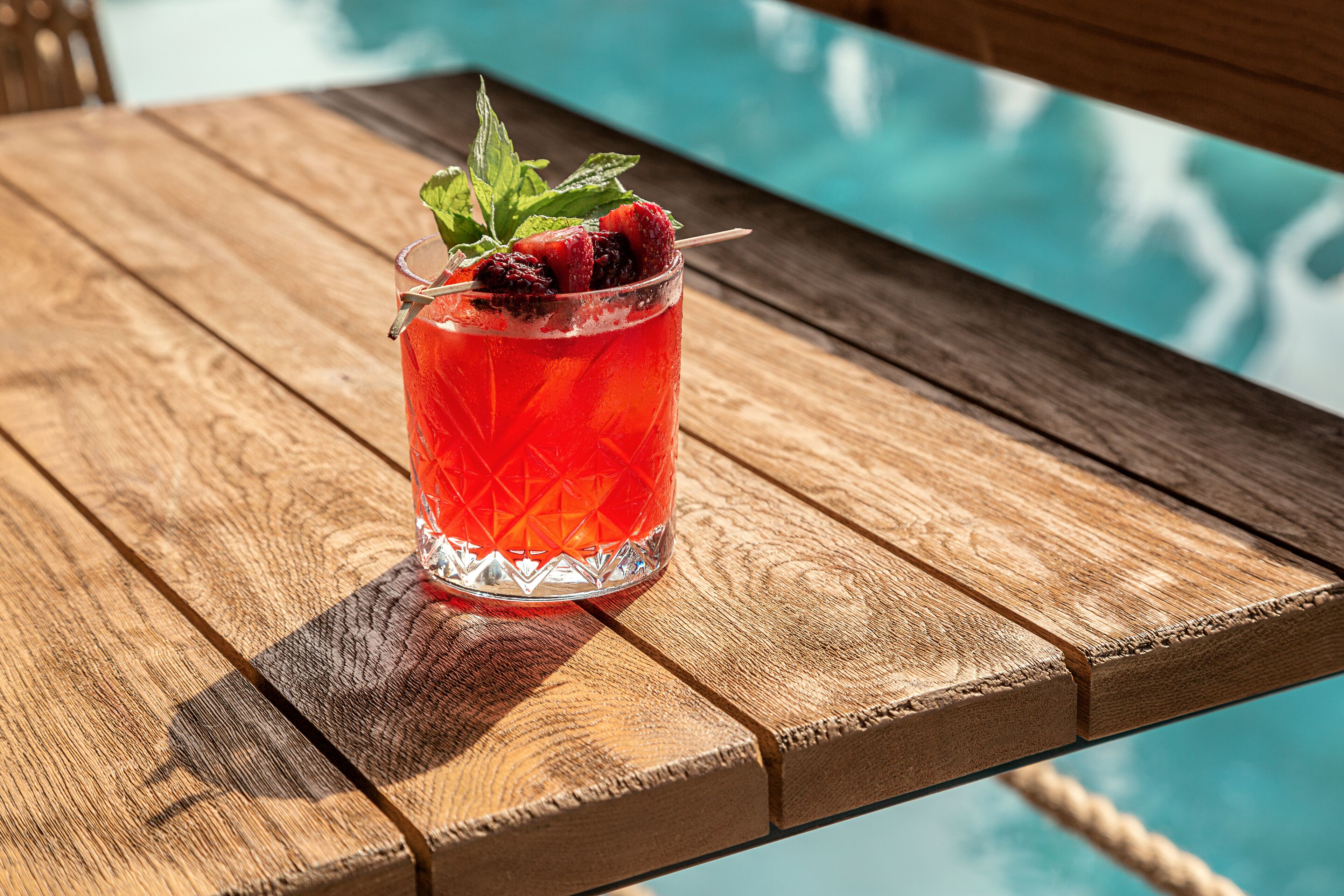 Un verre d’une boisson rouge aux fruits, posé sur une table en bois, près d’une piscine.