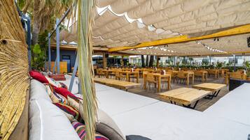 An outdoor seating area with wooden tables and chairs, a cushioned bench with colorful pillows, and a canopy providing shade.