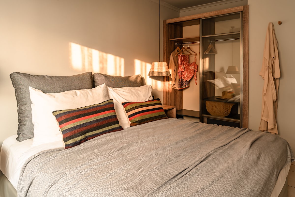 A neatly made bed with a grey and white patterned bedspread, a striped pillow, and a wooden nightstand with a lamp and a basket.