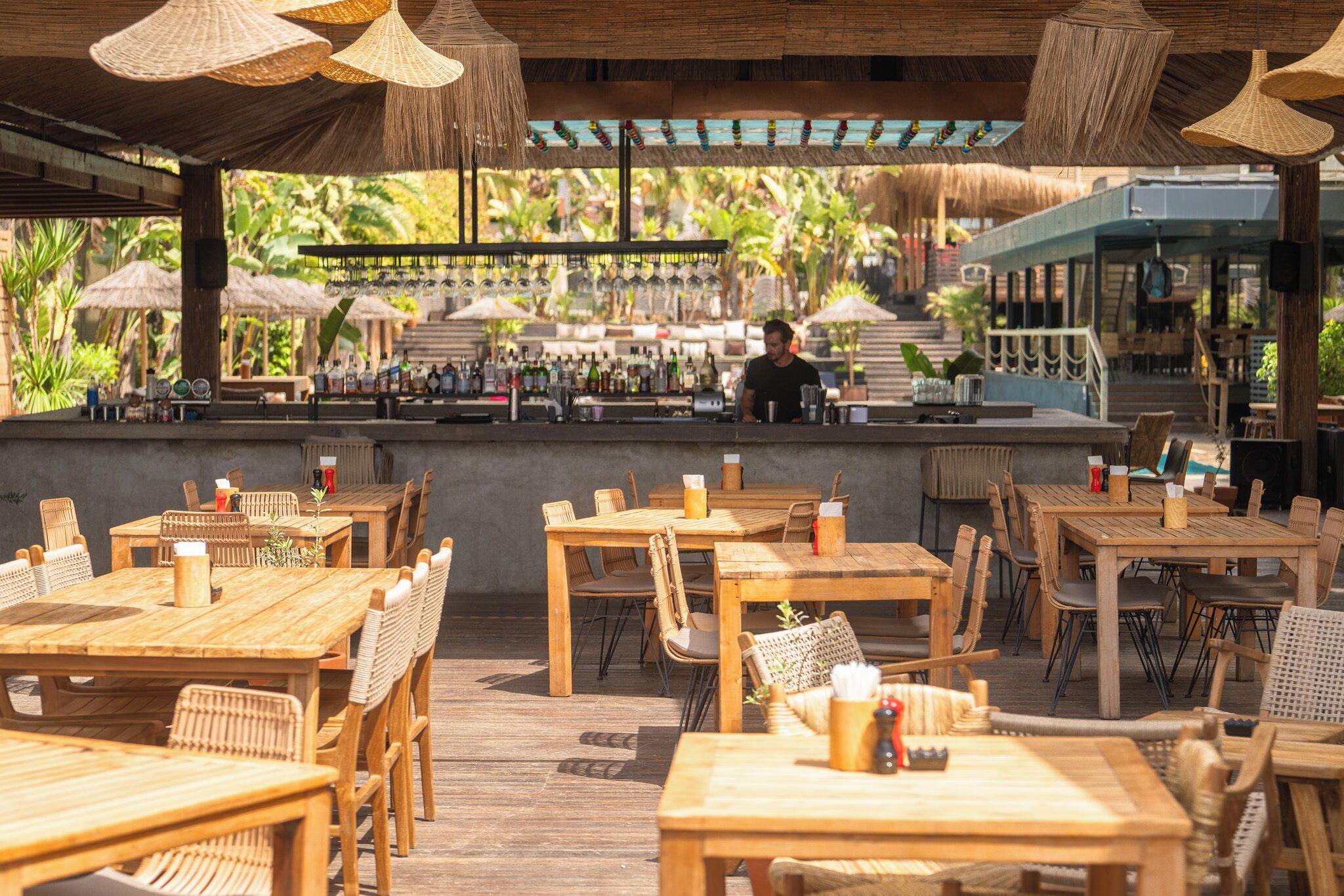 Un área de comedor al aire libre con mesas y sillas de madera, una barra al fondo y vistas a un jardín tropical.