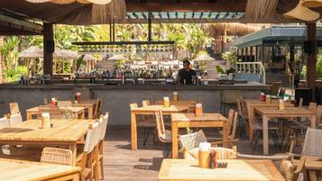 Un área de comedor al aire libre con mesas y sillas de madera, una barra al fondo y vistas a un jardín tropical.