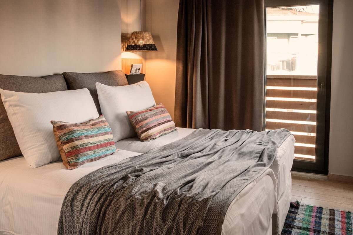 A neatly made bed with a gray blanket, two white pillows, and a colorful striped pillow. A small table with a lamp and a picture frame is beside the bed.