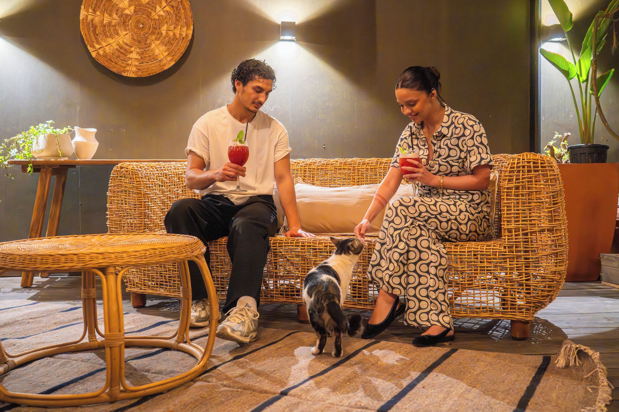 Two people seated on a wicker sofa with a small dog, a wicker table, and potted plants in a room with a textured wall and a round woven artwork.