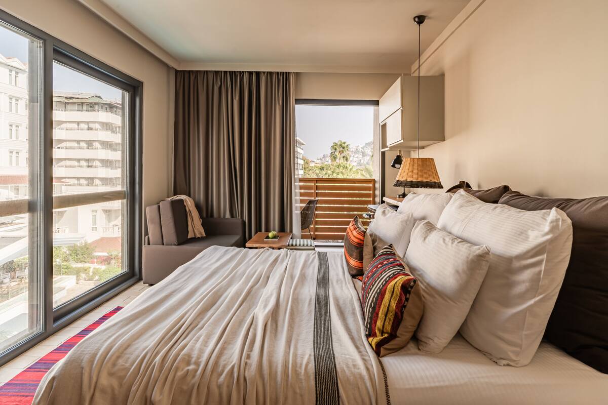 A hotel room with a large bed, a sofa, a desk, and a view of the city through large windows.