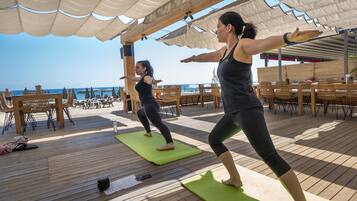 Deux personnes font de l’exercice sur des tapis verts, sous une terrasse en bois, avec vue sur la plage.