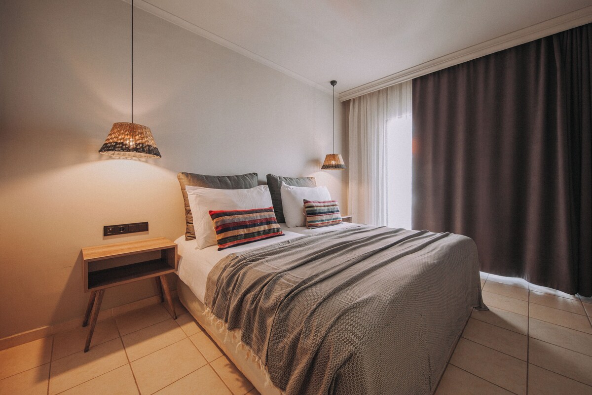 A neatly made bed with a patterned blanket and pillows, a bedside table with a lamp, and a view of a window with curtains.