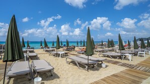 Een strand met ligbedden en parasols, een houten terras en een helder uitzicht op de oceaan.