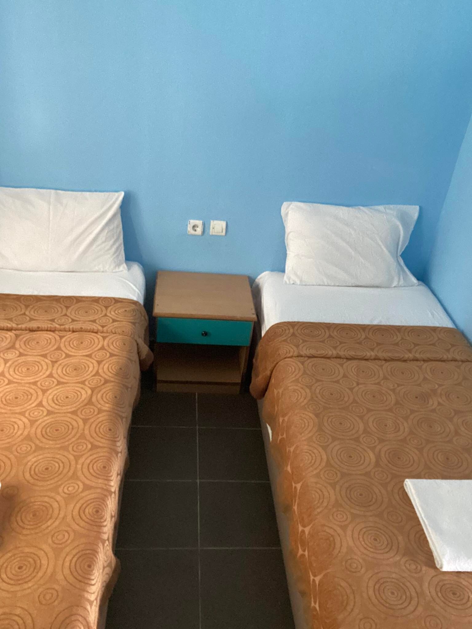 Ein Doppelzimmer mit zwei Betten, jeweils mit einem gemusterten Überwurf, einem kleinen Nachttisch und einer blauen Wand.
