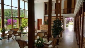 A spacious hotel lobby with high ceilings, large windows offering views of greenery, and a central reception desk.