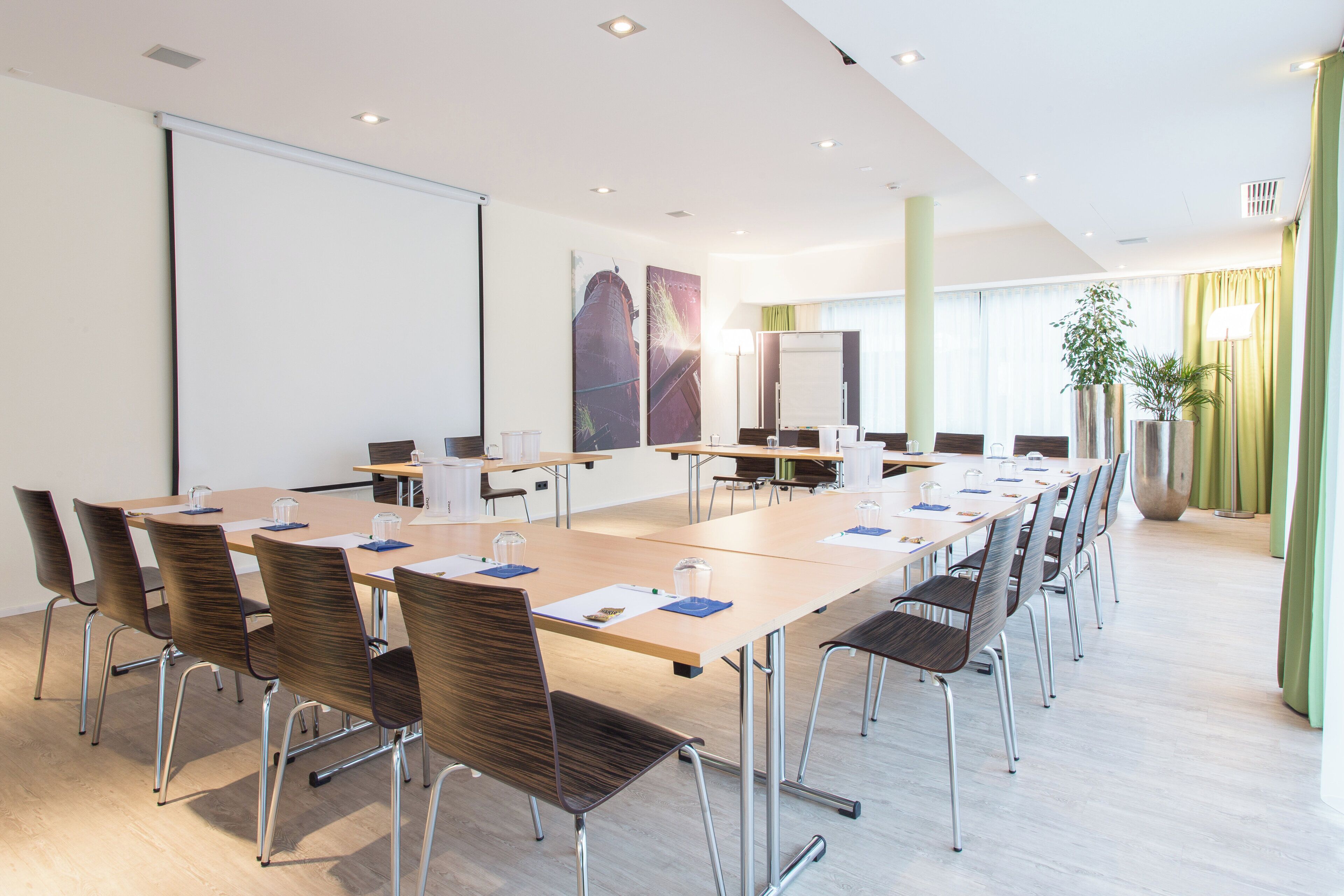 Ein Konferenzraum mit langem Tisch, Stühlen, einer Projektionsleinwand und einem großen Fenster mit Vorhängen.