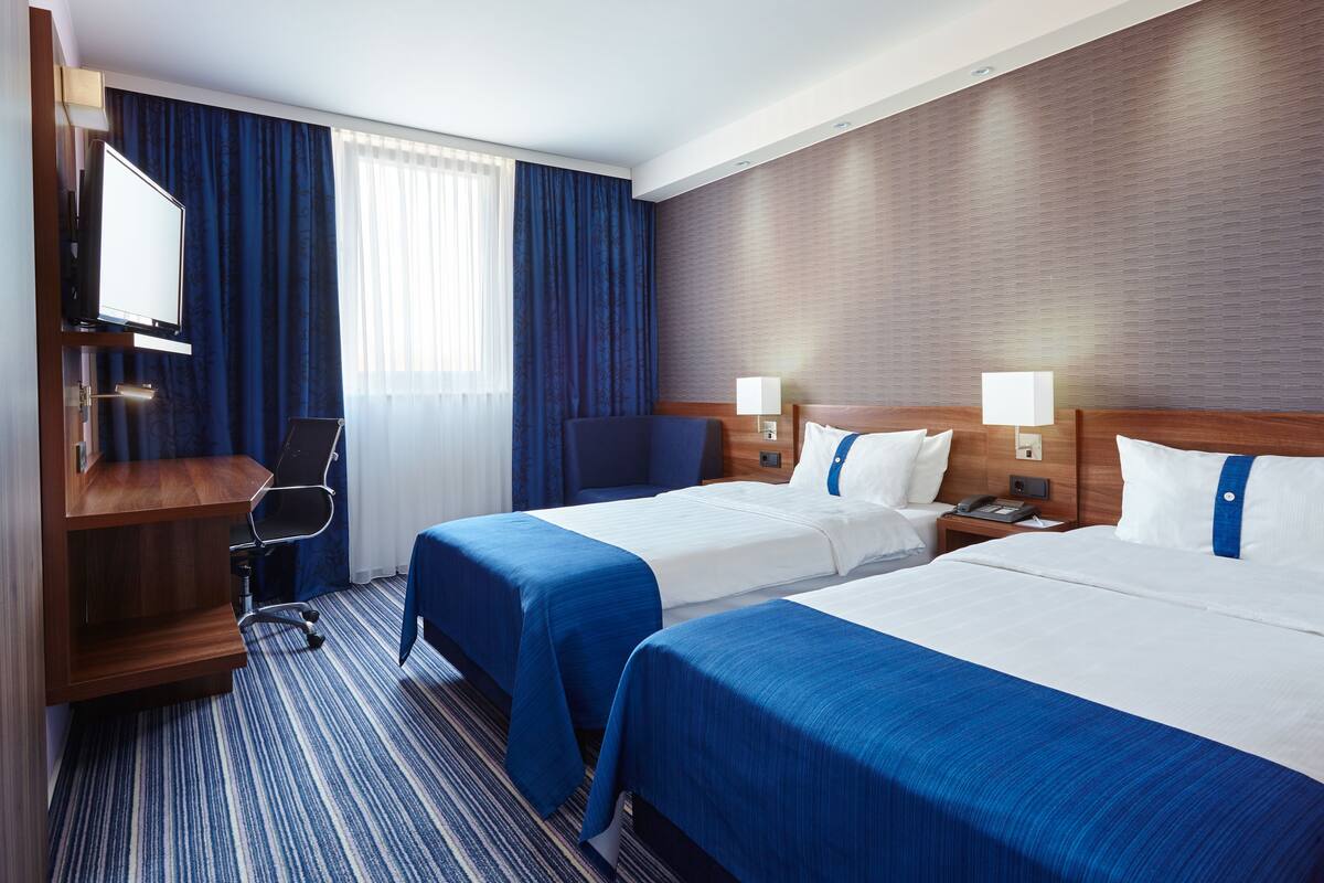 A hotel room with two beds, a desk, a chair, a TV, and a window with curtains.