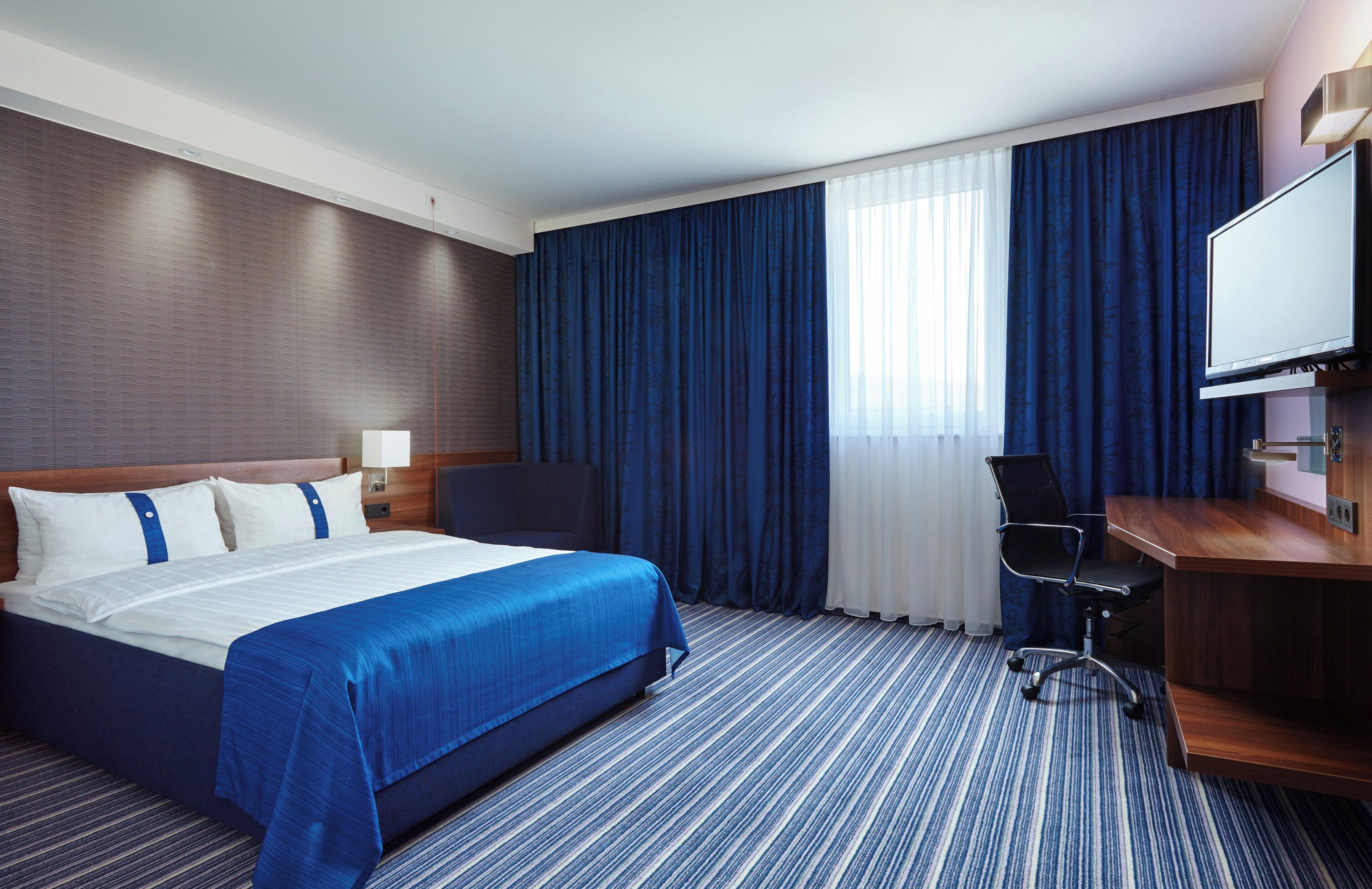 A hotel room with a large bed, a desk, a chair, a television, and a window with curtains.