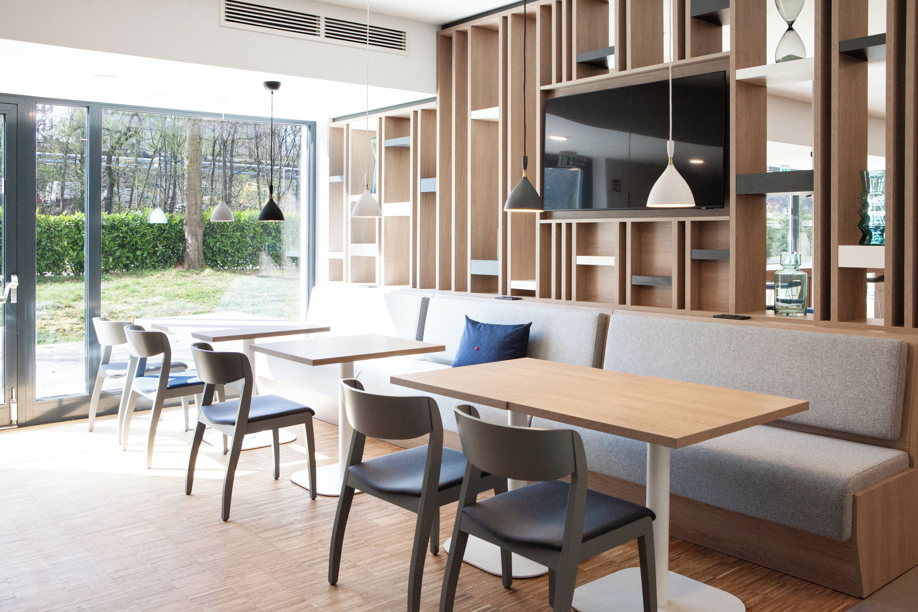 Ein modernes Interieur mit einer hölzernen Bank, Tischen und Stühlen, sowie einem großen Fenster mit Blick auf Grünflächen.