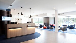 Un moderno hall de hotel con un mostrador de recepción de madera, múltiples lámparas colgantes y una zona de estar con sillas y sofás de colores.