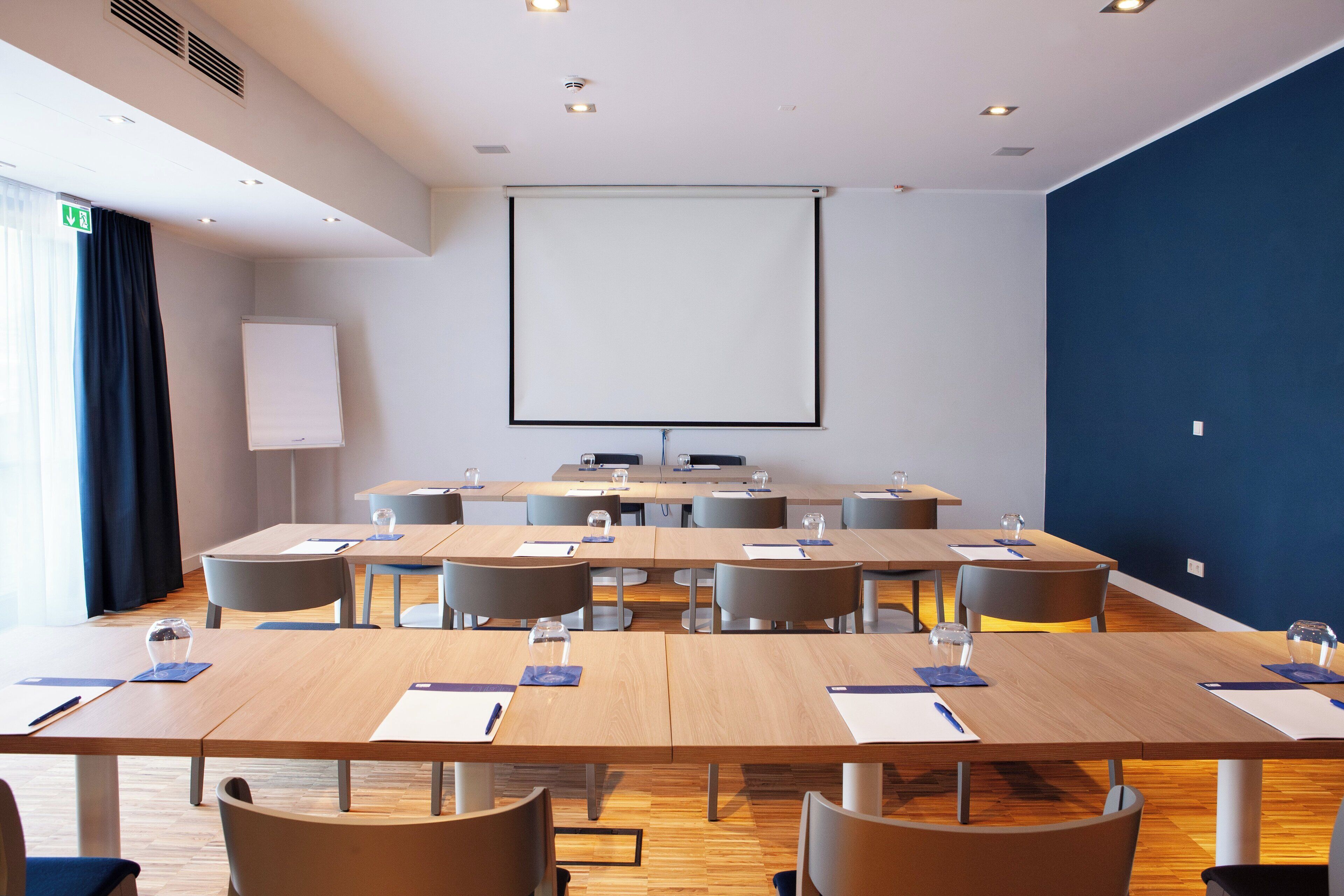 Ein Konferenzraum mit großem Bildschirm, mehreren Tischen und Stühlen.