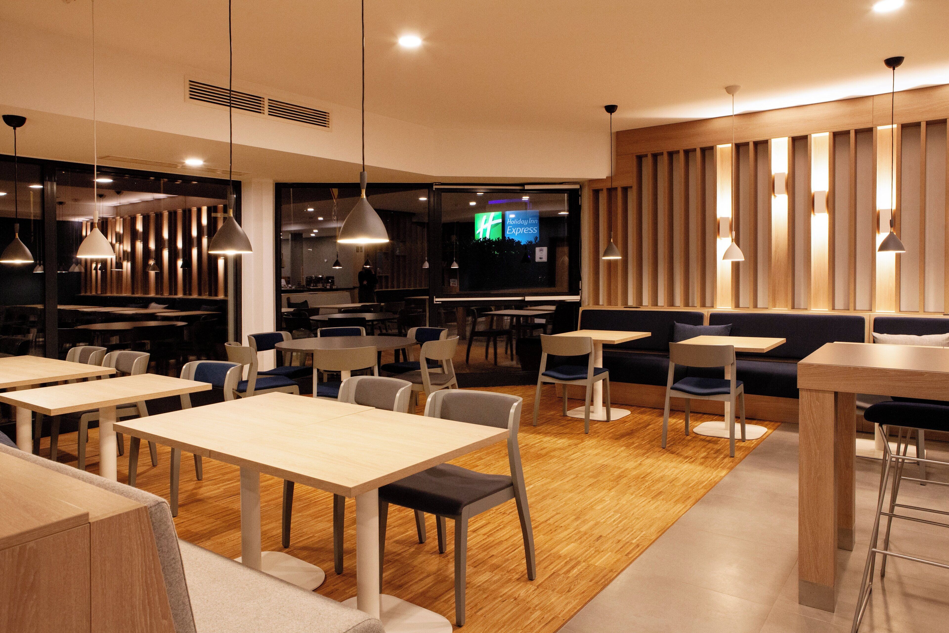 A modern restaurant with wooden tables and chairs, pendant lighting, and a view of the outside through large windows.